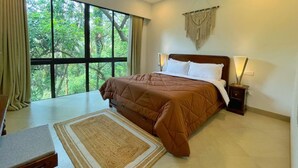A bedroom with a large window, a bed with a brown quilt, a nightstand with a lamp, and a ceiling fan. - Cozy 1BR in Candolim - Walking Distance to Beach (Candolim)