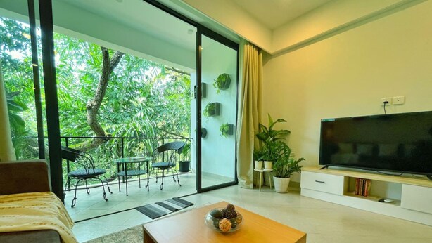 A modern living room with a sofa, coffee table, TV, and a balcony with chairs and plants. - Cozy 1BR in Candolim - Walking Distance to Beach (Candolim)