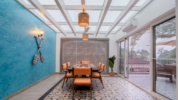 A dining area with a table set for six, a blue accent wall, and a large sliding glass door leading to an outdoor patio. - Irene Altezza by Hireavilla -2br w Pool in Pilerne (Marra)