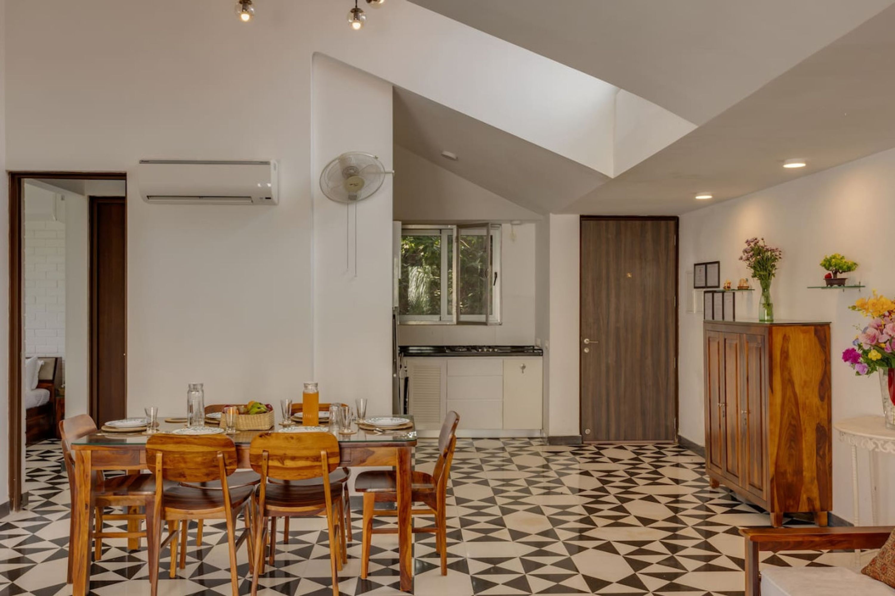 Un área de comedor con piso de estampado geométrico, una mesa de madera con sillas, una cocina al fondo y un armario de madera con un jarrón de flores.
