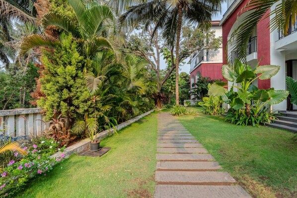A pathway leading to a building with a red facade, surrounded by lush greenery and palm trees. - 1BR Apt in Candolim With Pool 5 Mins From Beach (Candolim)