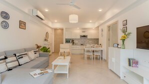 A modern living room with a grey sofa, white coffee table, and a dining area with a white table and chairs. - 1BR Apt in Candolim With Pool 5 Mins From Beach (Candolim)