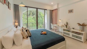 A modern hotel room with a large bed, a desk, and a balcony with a view of greenery. - 1BR Apt in Candolim With Pool 5 Mins From Beach (Candolim)