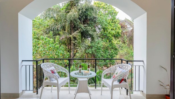 A balcony with white wicker chairs and a glass table, overlooking a lush garden. - 1BR Apt in Candolim With Pool 5 Mins From Beach (Candolim)