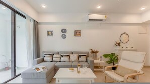 A modern living room with a grey sofa, a white coffee table, a wicker chair, and a large window with curtains. - 1BR Apt in Candolim With Pool 5 Mins From Beach (Candolim)
