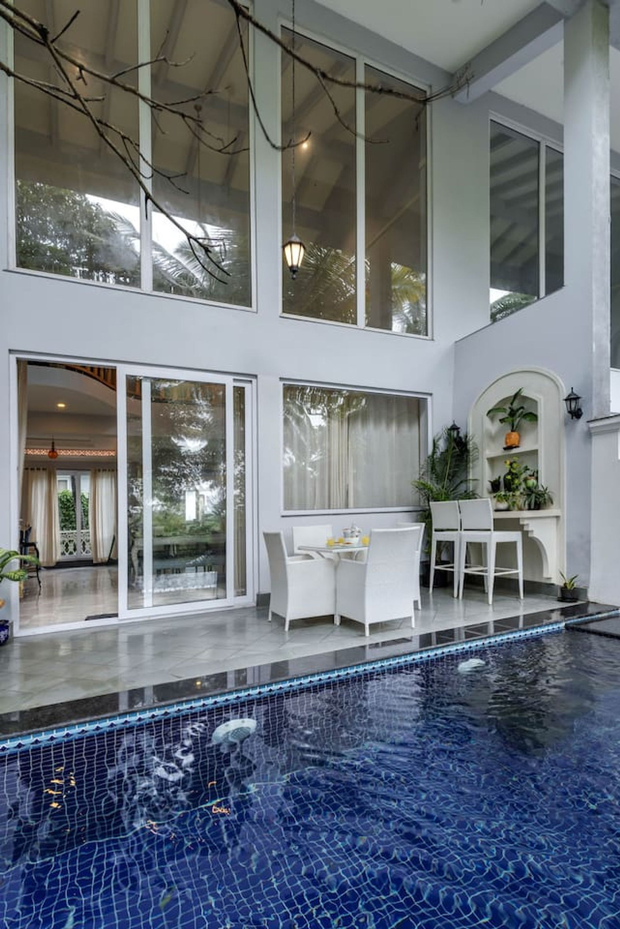 A modern indoor swimming pool with a glass enclosure, white furniture, and a view of the outdoors.