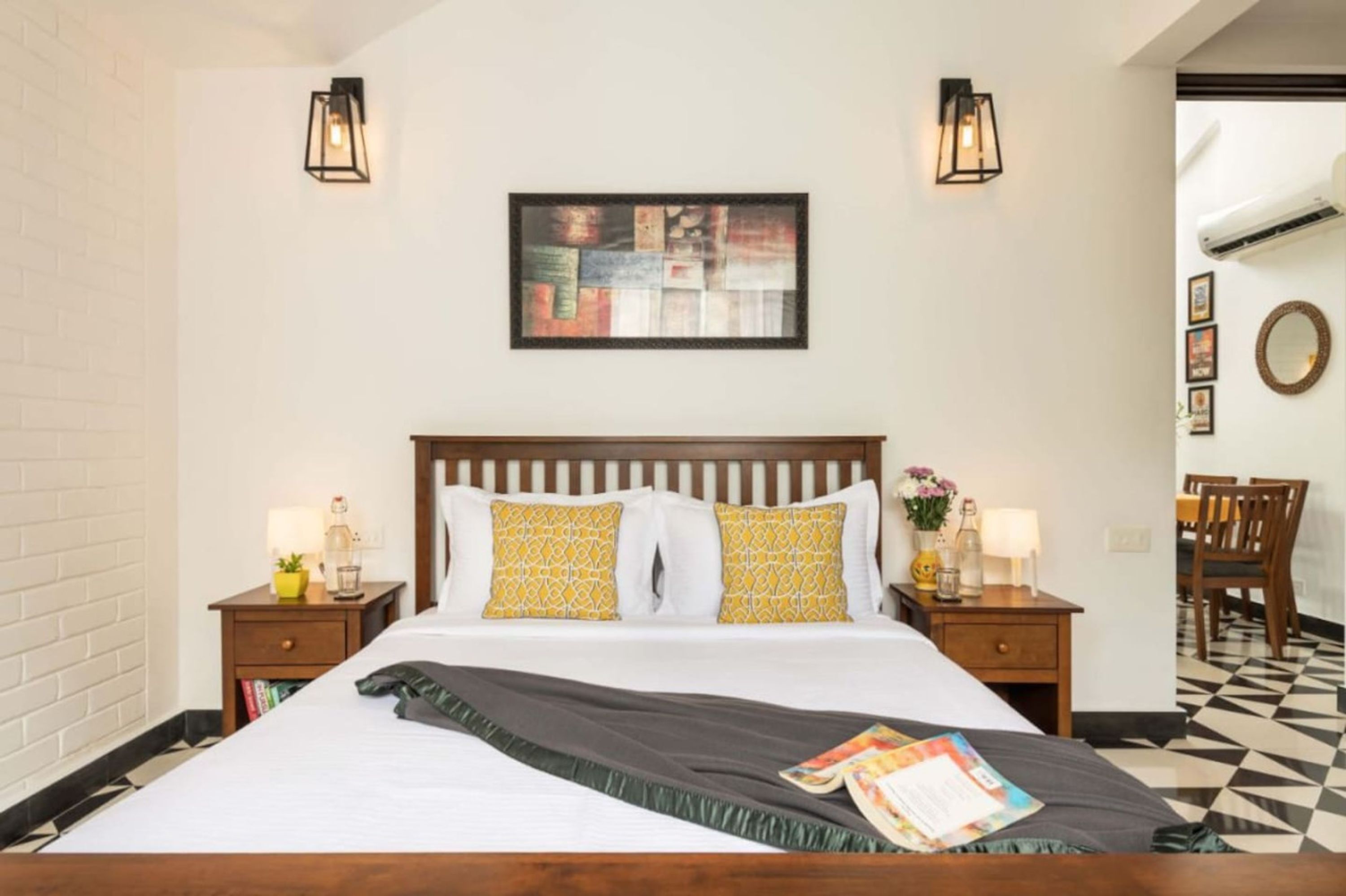 A hotel room with a wooden bed, two bedside tables with lamps, a wall-mounted artwork, a round mirror, and a checkered floor.