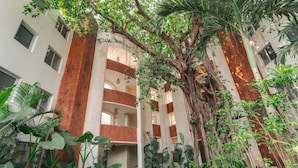 A multi-story building with a central atrium, surrounded by lush greenery and a large tree. - El Arbol by Hireavilla -2br Apartment in Nerul (Nerul)