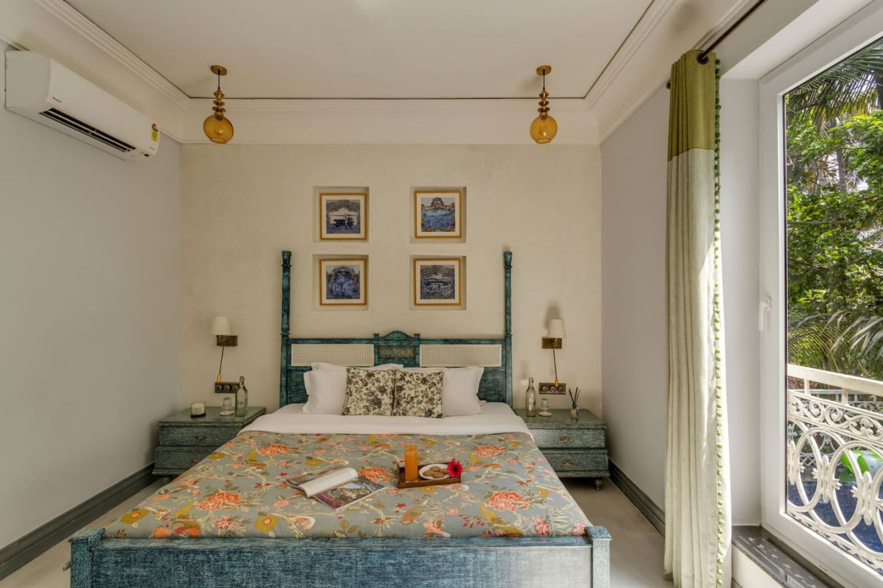 A bedroom with a bed, bedside tables, wall-mounted lamps, framed pictures, and a window with a view of greenery.