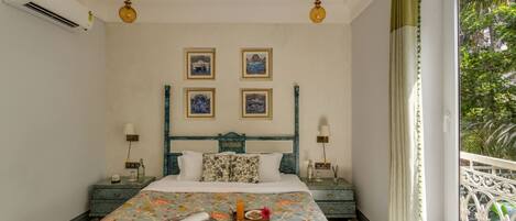 A bedroom with a bed, bedside tables, wall-mounted lamps, framed pictures, and a window with a view of greenery.