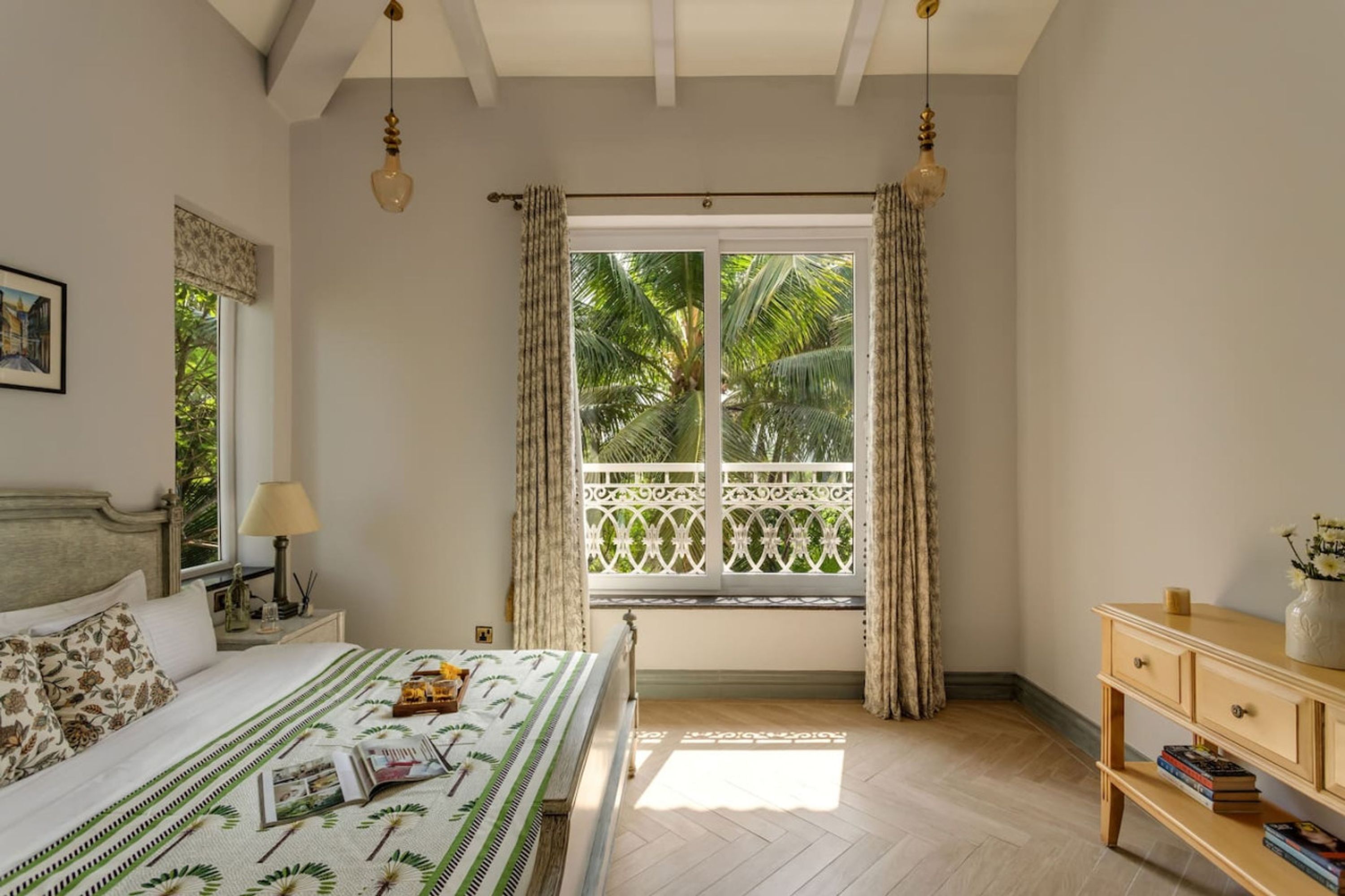 A bedroom with a bed, a nightstand, a lamp, a window with curtains, and a view of palm trees.