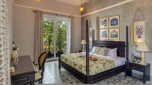 A four-poster bed with a floral bedspread, a nightstand with a lamp, and a view of greenery through a sliding glass door. - The Hornbill Villa - 3bhk With Pvt Pool in Saligao (Calangute)