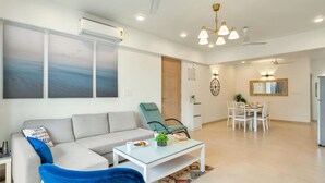 A modern living room with a sofa, a blue chair, a glass coffee table, and a dining area with a table and chairs. - Spacious 3BR Apartment With Pool in Candolim (Candolim)