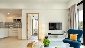 A modern living room with a glass coffee table, a blue armchair, a flat-screen TV, and a view of the kitchen and dining area. - Spacious 3BR Apartment With Pool in Candolim (Candolim)