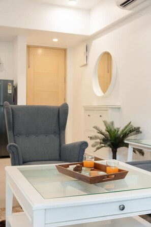 Een moderne woonkamer met een salontafel met glazen blad, een grijze fauteuil, een klein bijzettafeltje met een glas en een potplant.