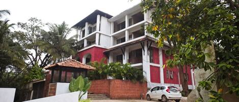 A two-story building with a red and white facade, a car parked in front, and a white wall with a small garden.