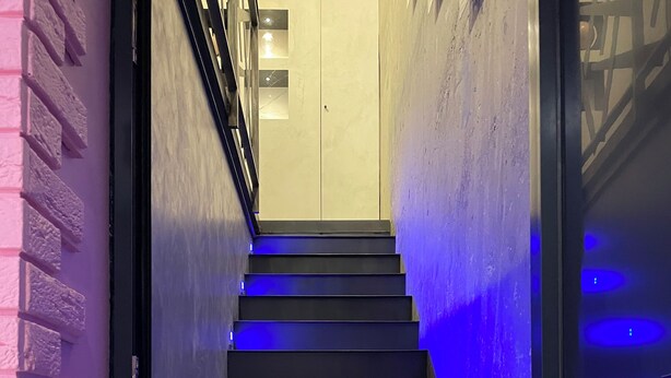 Eine Treppe mit blauer Beleuchtung, eine Ziegelwand links und eine Tür rechts.