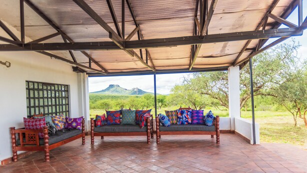 A covered outdoor seating area with two wooden benches, colorful cushions, and a view of green hills and mountains. - Ene Hacienda (Maili Tisa)