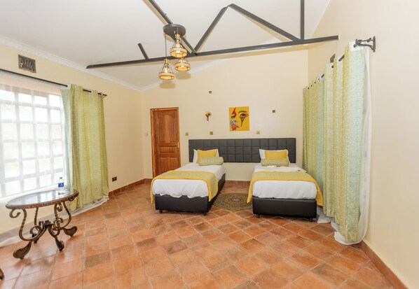 A room with two beds, a wooden door, a window with curtains, and a table with a decorative top. - Ene Hacienda (Maili Tisa)