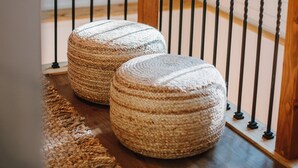 Una escalera de madera con barandilla de metal, dos otomanes tejidos y un tapete tejido sobre piso de madera.
