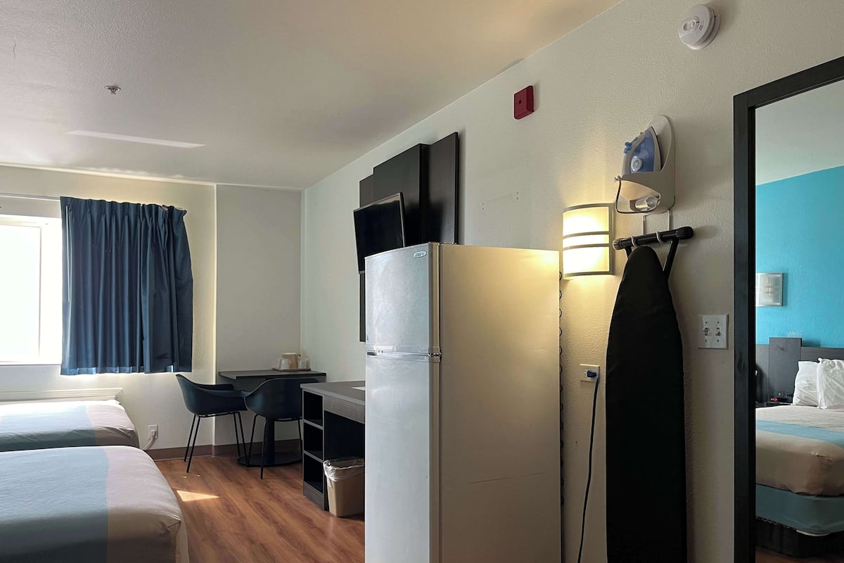 A hotel room with two beds, a refrigerator, a desk with a chair, a mirror, and a window with curtains.