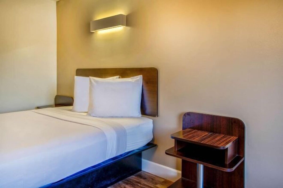 A hotel room with a bed, a nightstand, a wall-mounted light, and a wooden side table.