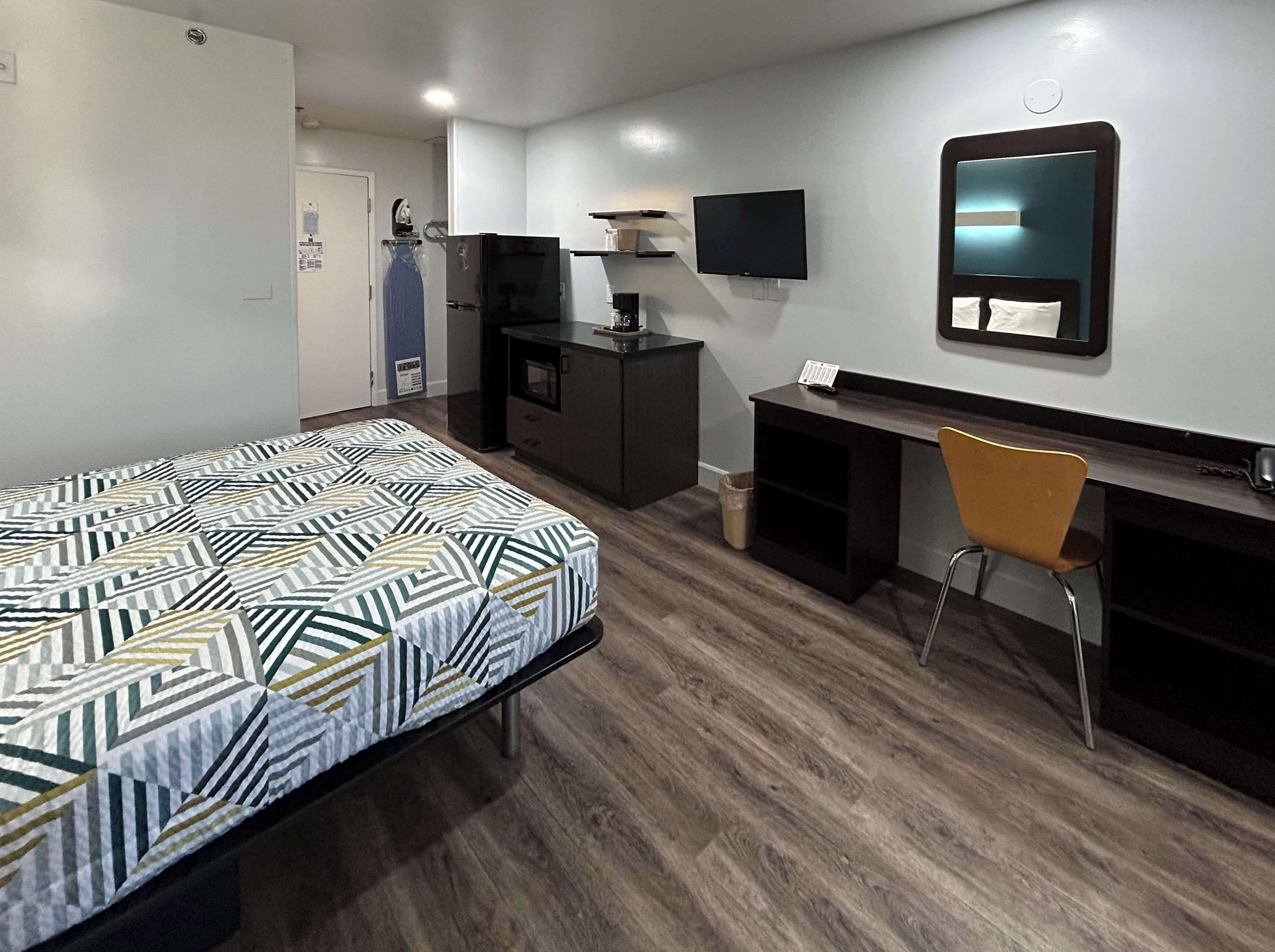 A compact hotel room with a bed, a desk, a chair, a TV, and a mirror.