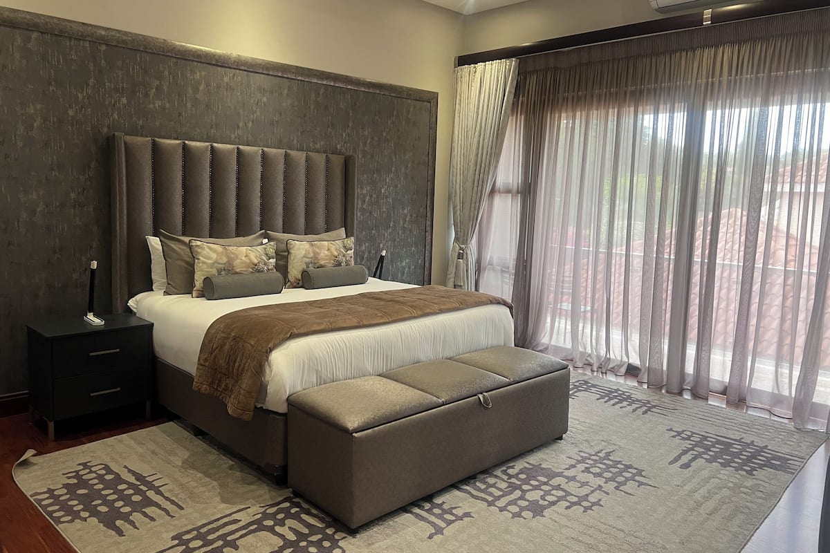 A modern bedroom with a large bed, a bedside table, a bench, and a view of the outdoors through sheer curtains.