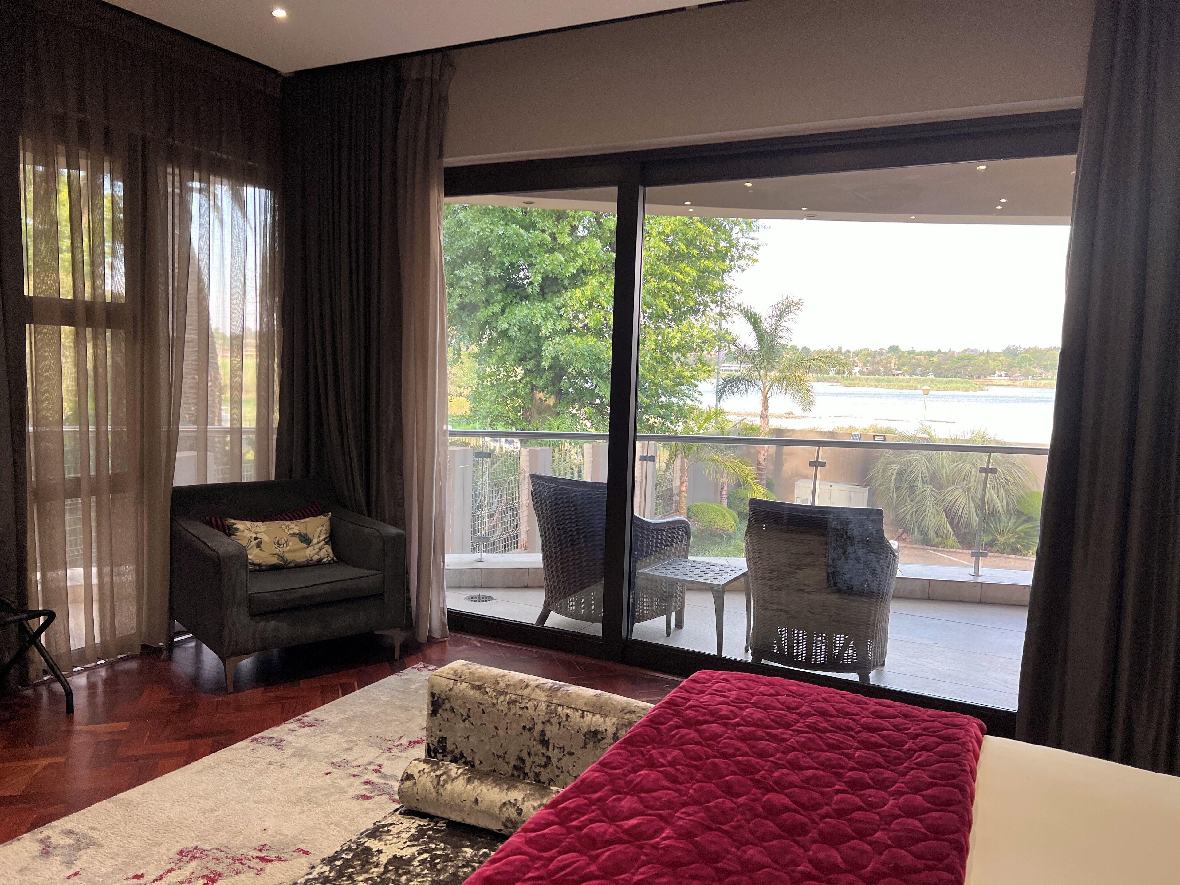 A modern room with a large sliding glass door leading to a balcony. The room features a sofa, a small armchair, and a bed with a red blanket.