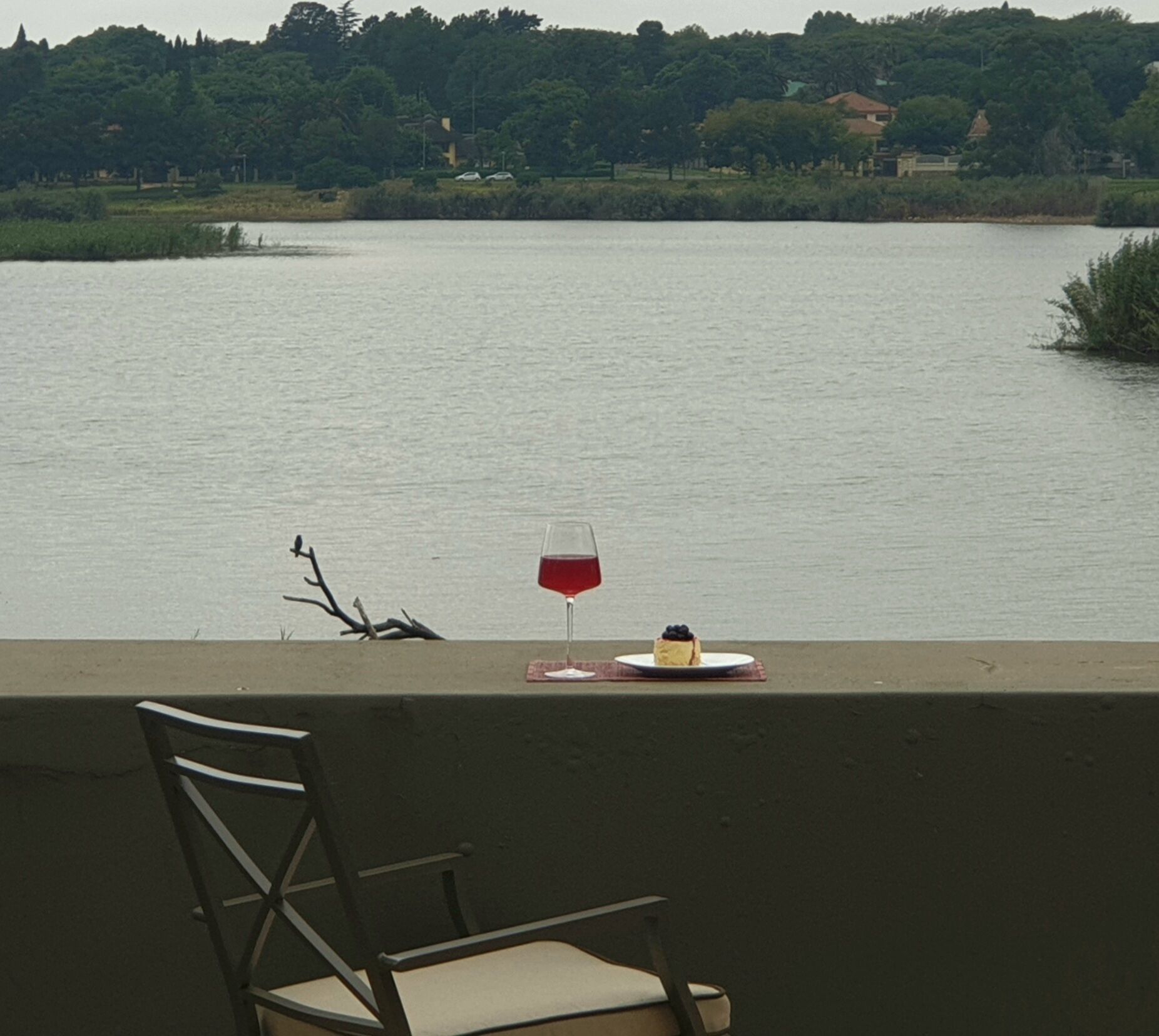 Una silla y una mesa con una copa de vino tinto y un postre junto a un lago.
