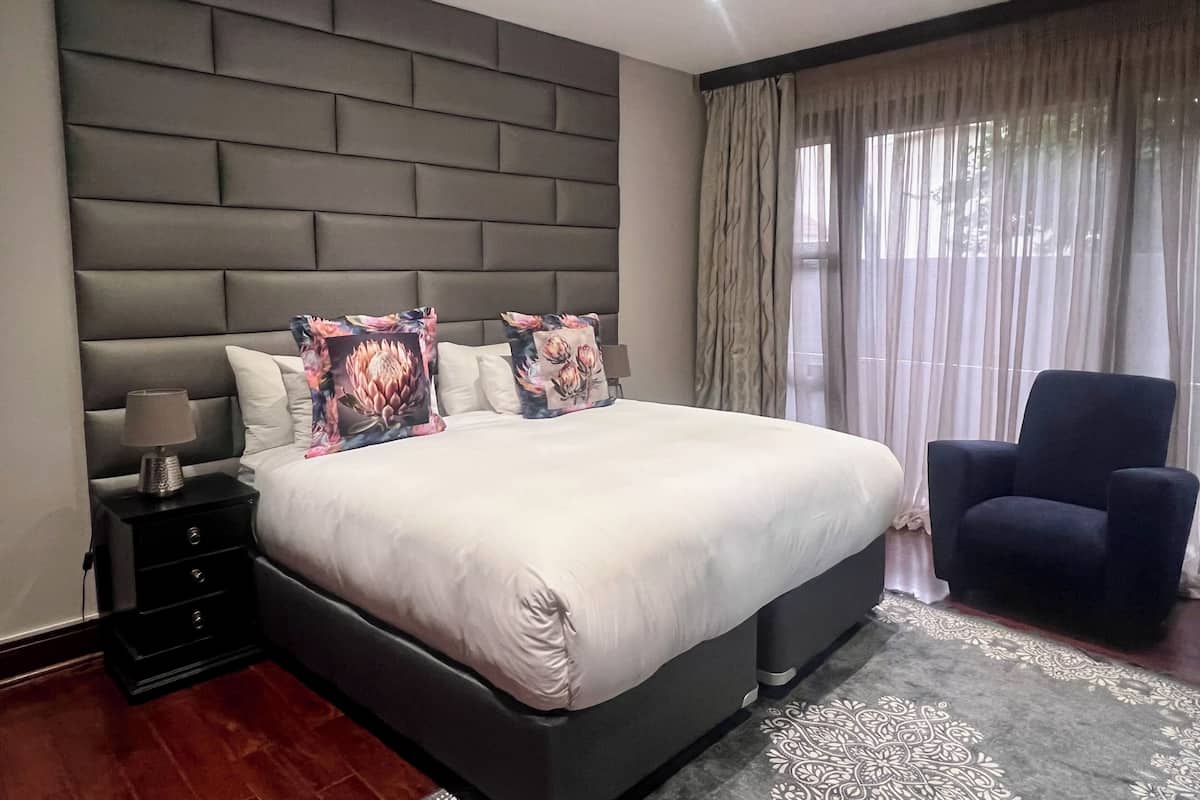 A modern bedroom with a large bed, a nightstand, a lamp, a chair, and a view of the outdoors through sheer curtains.