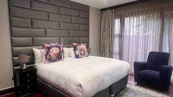 A modern bedroom with a large bed, a nightstand, a lamp, a chair, and a view of the outdoors through sheer curtains.
