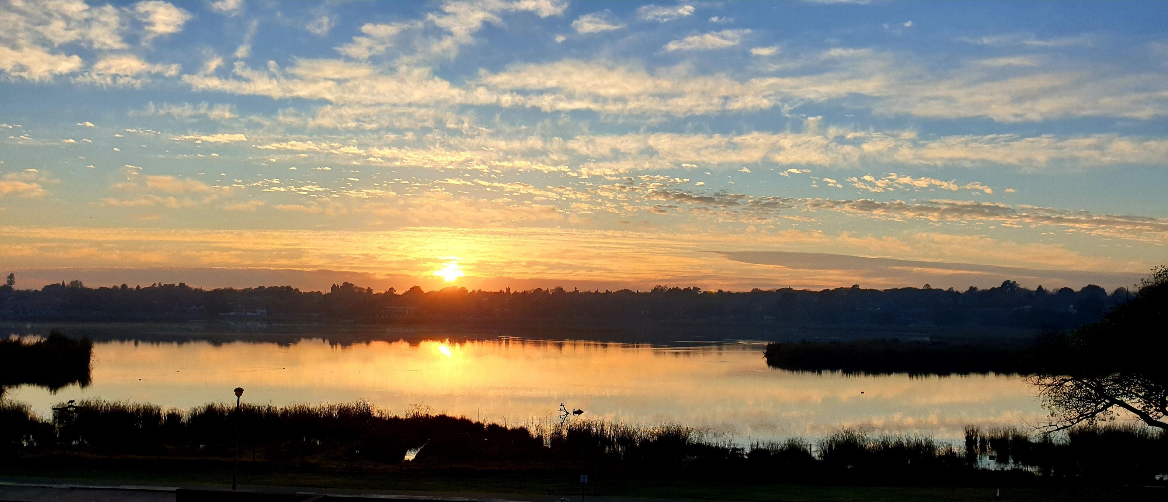 Un coucher de soleil sur un lac paisible, avec la silhouette d’arbres et, au loin, une ville.