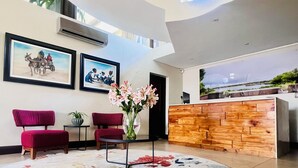 Un vestíbulo moderno con una recepción, dos obras de arte enmarcadas, un sillón tapizado rojo, una pequeña mesa con un jarrón de flores y una alfombra colorida.