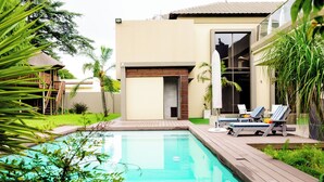 Un área de piscina moderna con un deck de madera, sillas de descanso y una puerta de vidrio que da a un porche cubierto.