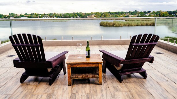 Dos sillas Adirondack, una mesita de madera y una botella de vino en una terraza con vistas a un cuerpo de agua.