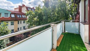 Ein Balkon mit grünem Kunstrasen, Metallgeländer und Blick auf benachbarte Gebäude und Bäume.