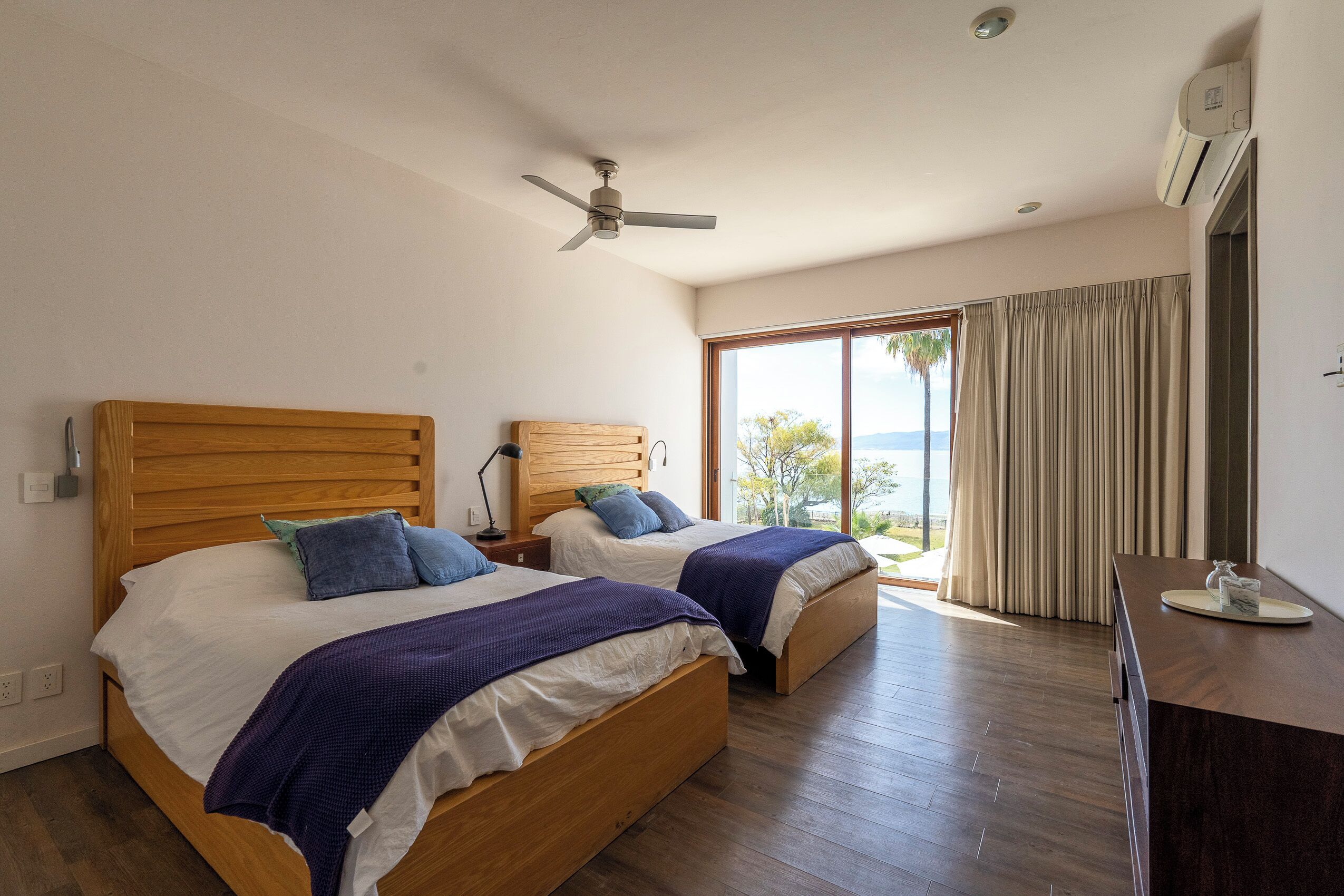 A hotel room with two wooden beds, a ceiling fan, an air conditioning unit, a large window with curtains, and a wooden desk with a mirror.
