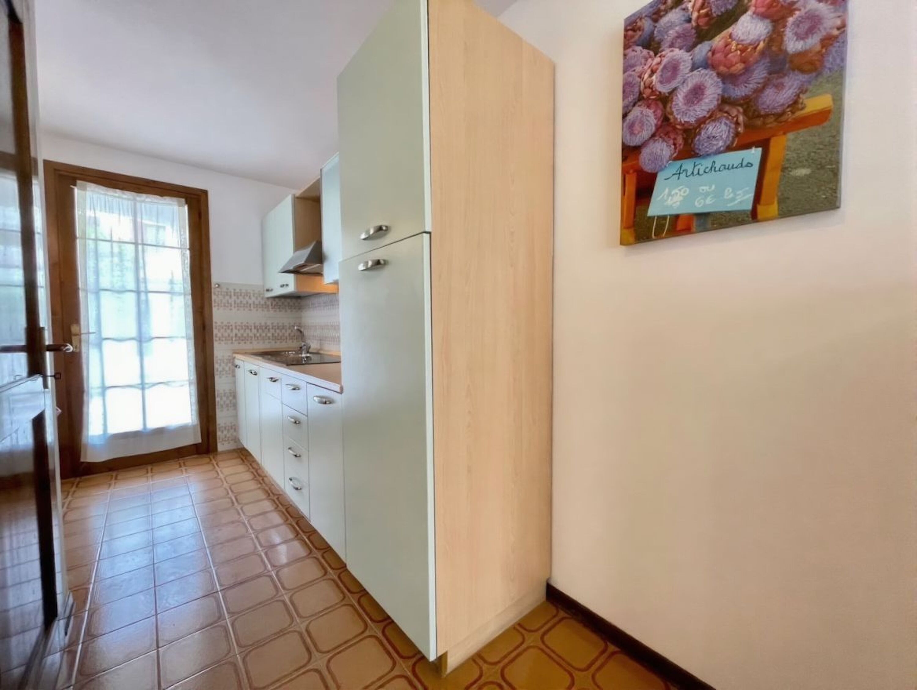 A kitchen with white cabinets, a sink, and a stove. There is a door leading to another room and a window with curtains.