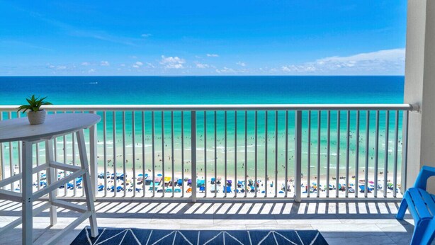 A balcony with a view of a beach, a table with a plant, and blue chairs.