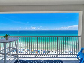 A balcony with a view of a beach, a table with a plant, and blue chairs.