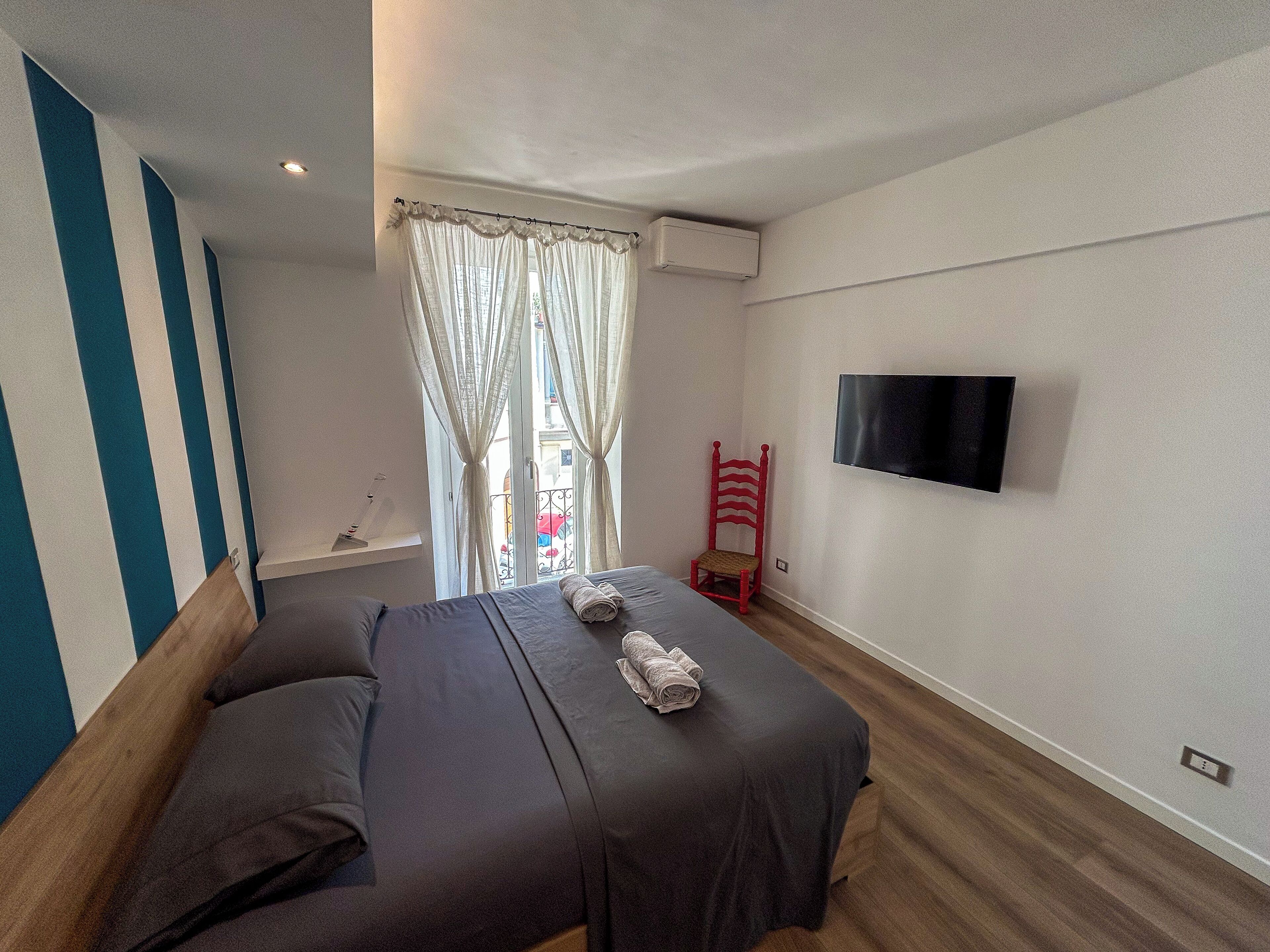 Ein Schlafzimmer mit einem Bett, einem roten Sessel, einem Fernseher und einem Fenster mit Vorhängen.