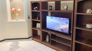 A living room with a flat-screen TV mounted on a wooden entertainment unit, a small shelf with decorative items, and a vase with flowers on a window sill. - New Apt in GDL Near Cathedral  (Guadalajara)