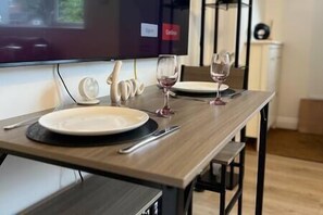 A dining table set with plates, cutlery, and wine glasses, with a television mounted on the wall behind.