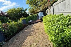 A garden pathway leading to a white door, surrounded by lush greenery and a wooden fence.