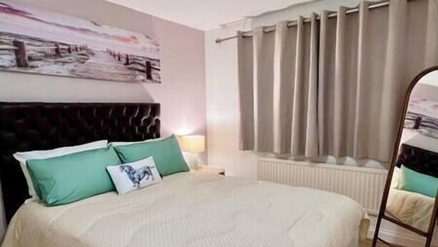 A bedroom with a bed, pillows, a headboard, a nightstand, a lamp, a window with curtains, and a radiator.