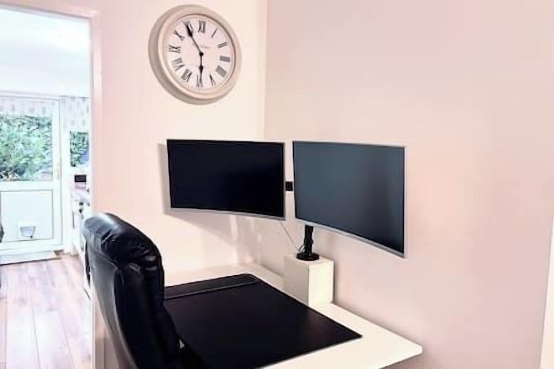 A room with a bed, a desk, two monitors, a clock, and a window.