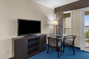 Ein Hotelzimmer mit Flachbildfernseher auf einem Holzschrank, einem runden Tisch mit zwei Stühlen und Blick auf den Balkon durch eine Tür.
