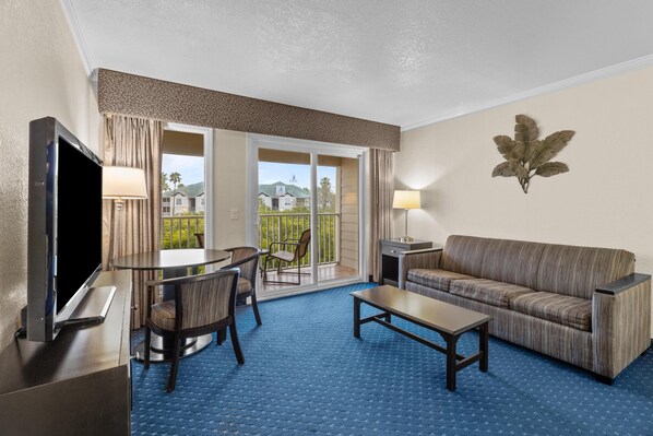 A hotel room with a flat-screen TV, a sofa, a coffee table, a dining table with chairs, and a balcony with a view of houses.