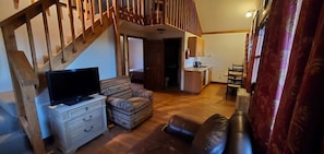 Una sala de estar con escalera de madera, televisor sobre una cómoda, un sofá y una zona de cocina al fondo.